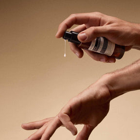 A close-up of a person's hands holding and spraying an amber glass bottle with a black pump dispenser, with a single drop of Aesop Solais Replenishing Hand Serum dripping down, against a warm beige background.