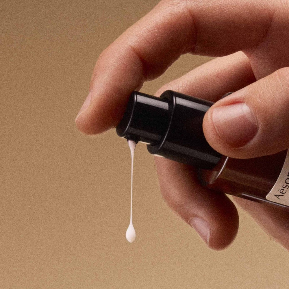 A close-up of a hand holding a dark pump bottle and dispensing a single drop of white cream against a warm beige background.