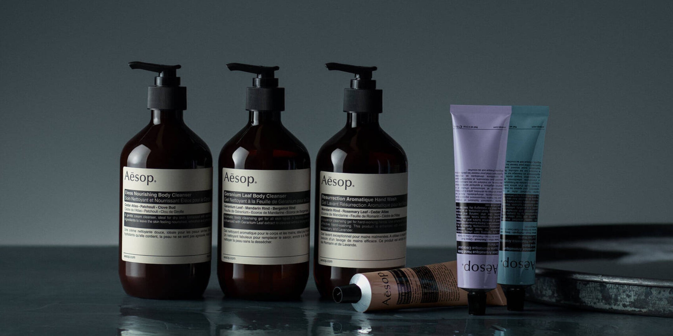 Four Aesop skincare and body care products arranged on a dark reflective surface against a dark gray background: three amber pump bottles with cream labels and two squeeze tubes, one lavender and one blue, with one tube lying on its side.