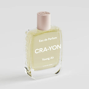 A clear glass bottle of Cra-Yon Young Air Eau de Parfum with pale yellow liquid, a light pink cap, and white lettering, set against a clean light gray background.